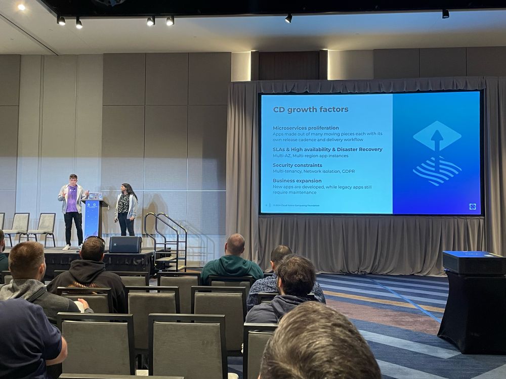 Picture of KubeCon stage with Pinky & Leigh standing at the speaker podium and slide share screen with a title of “CD growth Factors” with key points. 