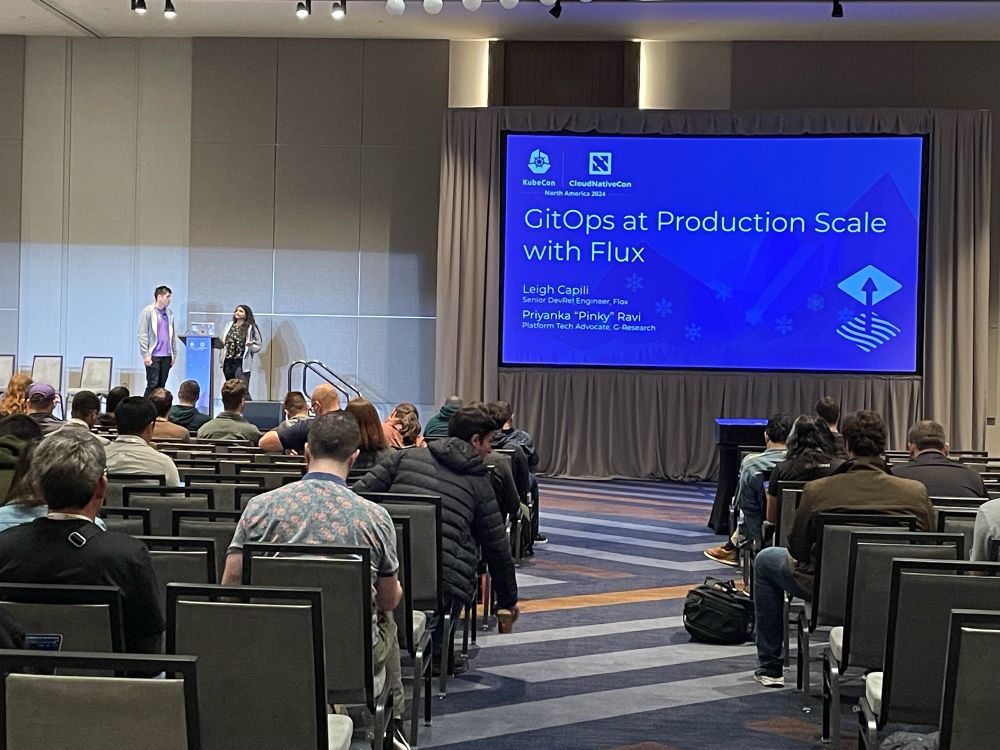 Picture of KubeCon stage with Pinky & Leigh standing at the speaker podium and screen of slide share with GitOps at Production Scale with Flux in white in a blue background. 