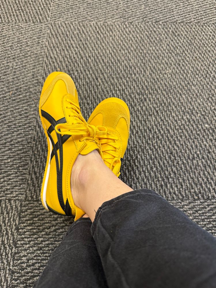 A pair of women’s legs in black jeans crossed at the ankles. She is wearing yellow Onitsuka Tiger Mexico 66 sneakers. 