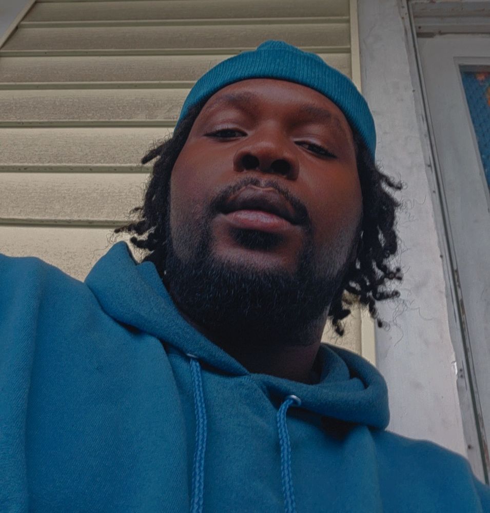 A from the bottom up selfie of the user, he has short locs, a short beard, and is donned in a blue brimless hat and blue hoodie 

He sits in front of a very light green wall with a white doorframe in frame