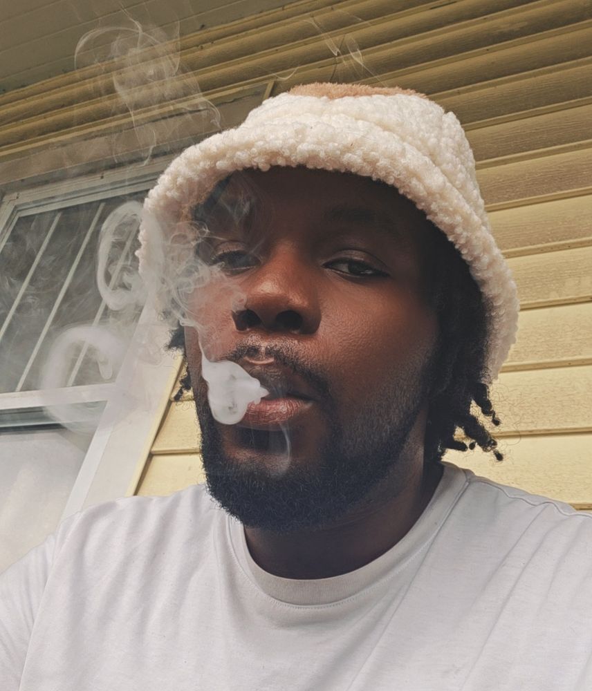 A selfie of the user, he sports a short beard and short locs and dark skin. He is adorned in a fleece bucket hap styled after a sky bison from the series "Avatar: The Last Airbender" and a simple white tee, he sits Infront of a window and yellow green wall