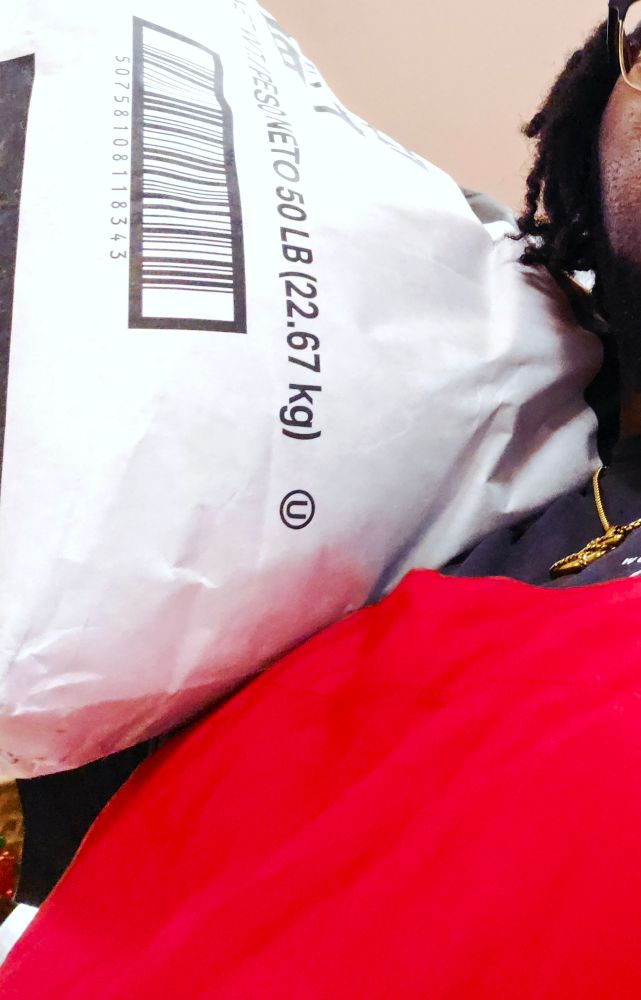 An upwards faceless selfie of the user where in one arm they hold a 50lb sack of sugar on their shoulder, they have on a black shirt with a red apron, simple gold rope chain with an anchor pendant, and some locs creep in from the right

The sack is flour white and reads as being 50lbs
