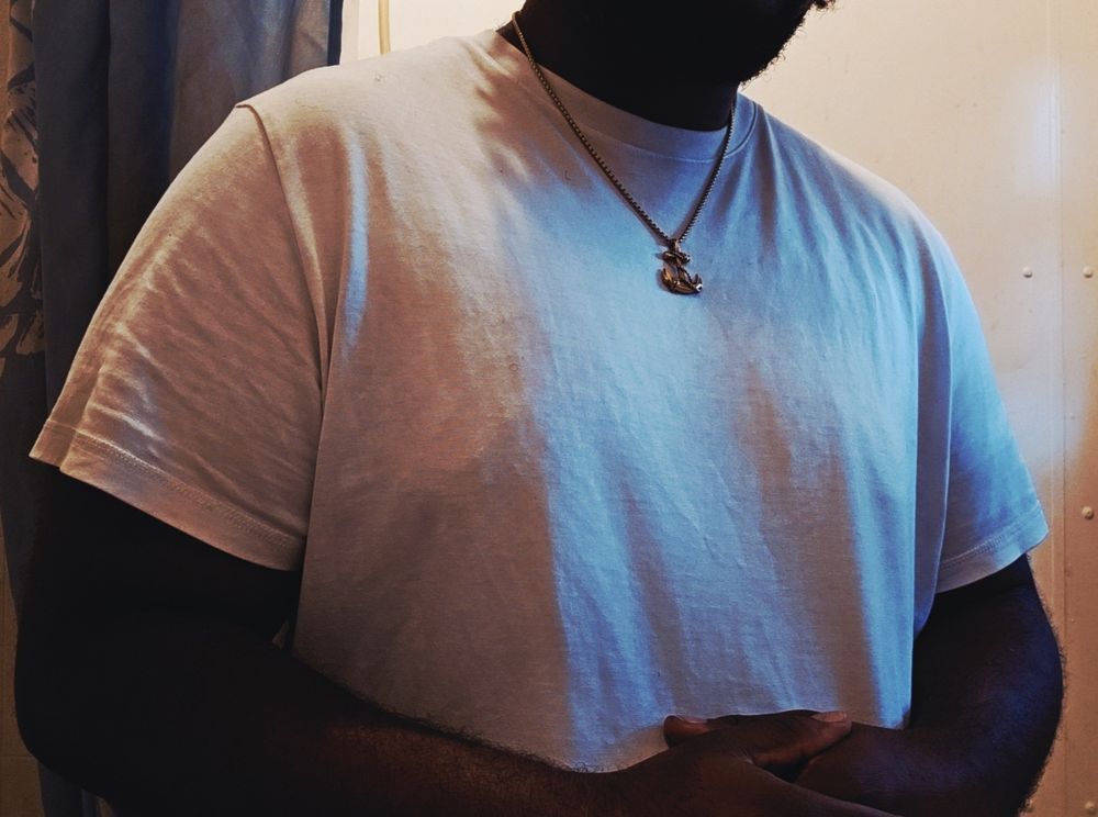 A selfie of the user from the jaw down but above the waist, he is donned in a white t shirt with a simple gold rope chain with an anchor pendant. His hands rest on top of each other at his natural waiste, he stands amongst a white wall with a light blue shower curtain behind him