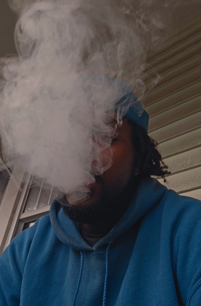 A from the bottom up selfie of the user, he has short locs, a short beard, and is donned in a blue brimless hat and blue hoodie 

He sits in front of a very light green wall with smoke obscuring most of his visage 