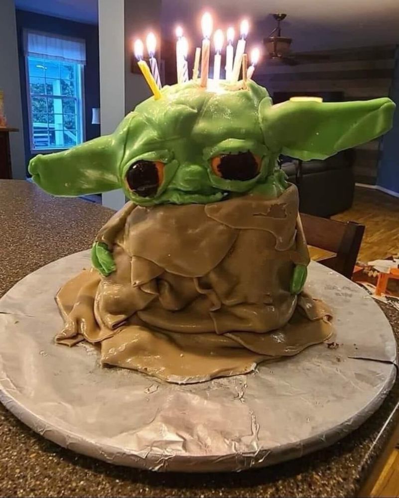 A Yoda shaped cake that looks like it's melting and has leprosy with orangeish red eyes and birthday candles on its head 