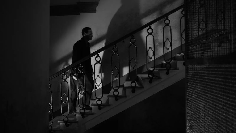Ripley ascending a staircase, the walls frame him like an uneven triptych and his shadow looms enormously in front of him on the wall 