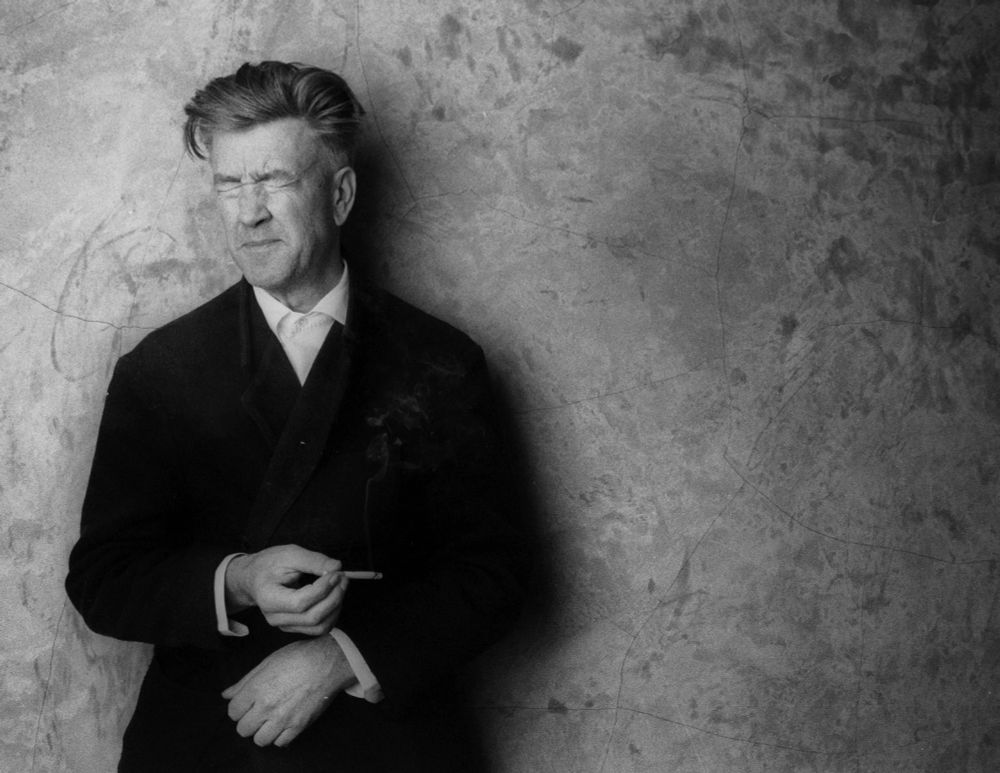 David Lynch, squinting, holding a cigarette weirdly between his fingers while standing against a cement wall.