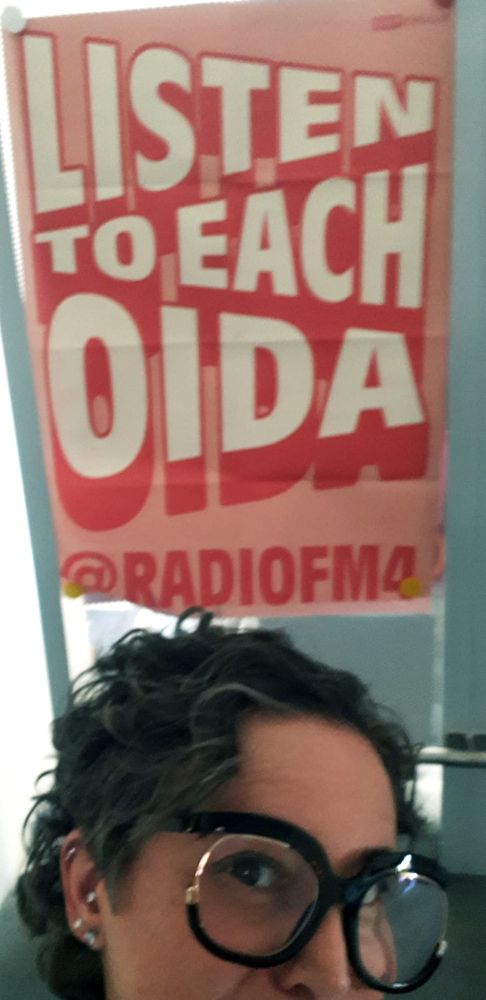 a selfie of me in the Radio FM4 office with a poster that says "Listen to each oida" behind me and I am wearing my humongous round black glasses -Iris Apfel Forever- and my hair is short and curly and very cute and I have diamond earring studs in my right ear. not real. 