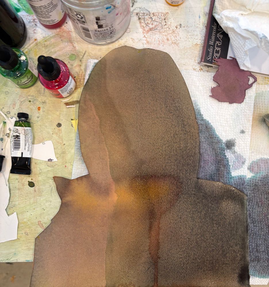 picture of a tabletop showing bottles and tubes of paint and a brownish gray piece of paper cut in the silhouette of a man holding a cat 