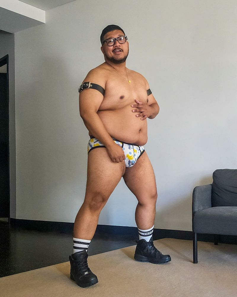 Chunky brown man wears black leather armbands and boots, a small rubber-duck necklace, and rubber-duck themed underwear.