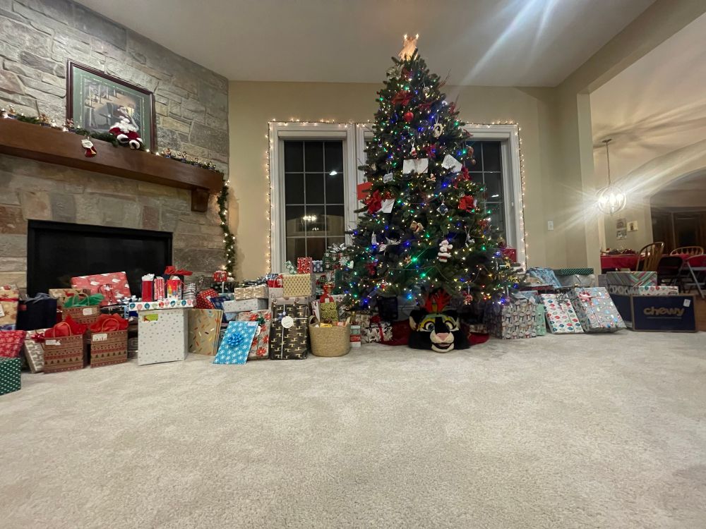 Brightly lit Christmas tree surrounded by presents with a fursuit head hidden underneath 