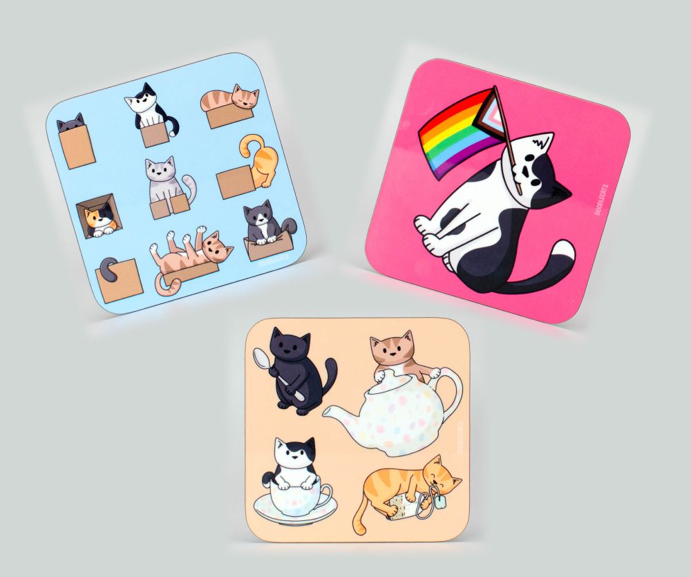 Three coasters. One is blue with nine cats in boxes doing various silly things. One is pink and has a black and white cat holding a progress pride flag in their mouth. the third is a beige coaster with four cats, one with a tea spoon, one with a teapot, one with a tea cup and the last with a tea bag