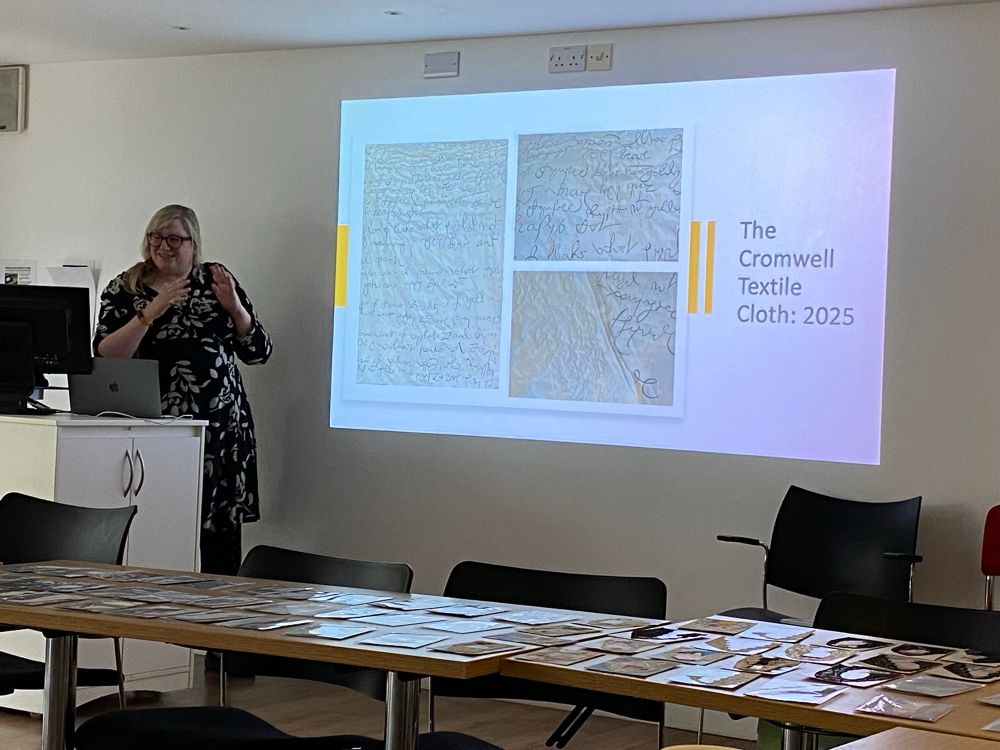A white woman wearing a black and cream dress stands beside a presentation screen that shows images of a quilted piece of artwork. The screen reads “The Cromwell Textile Cloth 2025”. 