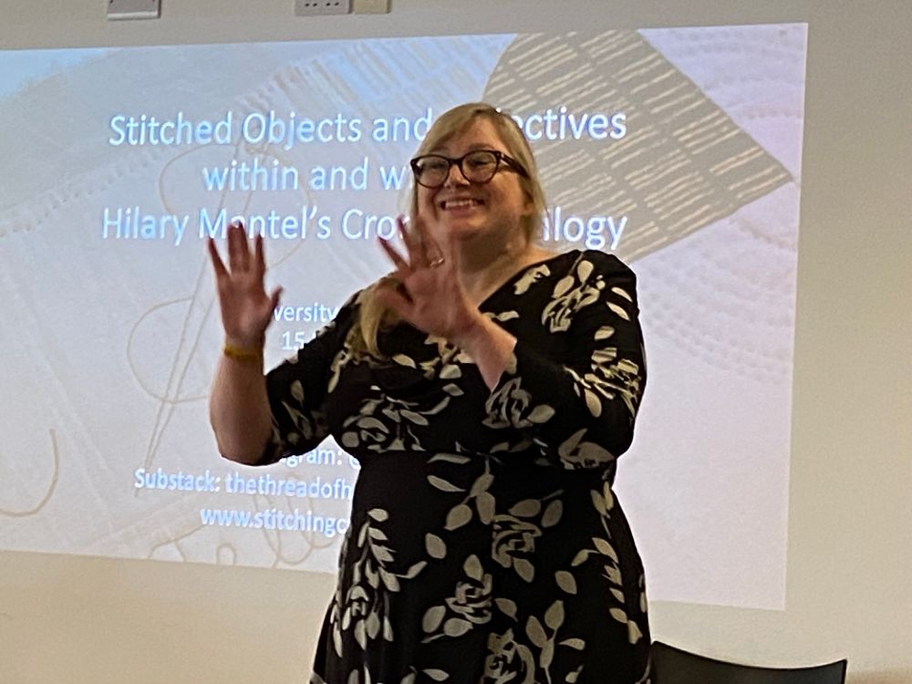 A white woman wearing a black and cream dress stands in front of a presentation screen. 