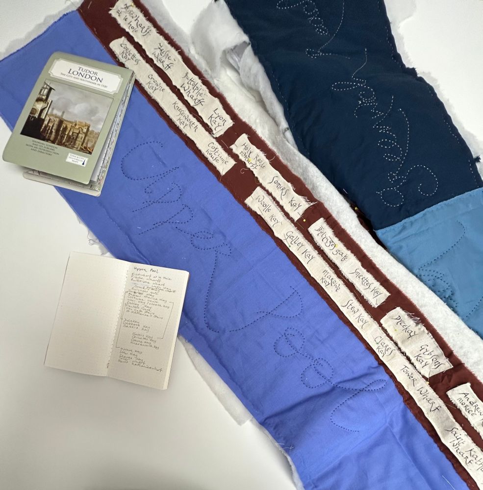 A map of Tudor London, folded. A notebook with Thames-related notes. A quilted piece of blue fabric, with the words Upper Pool stitched; then a strip of brown fabric with the names of different wharves and quays inked on cream calico. 