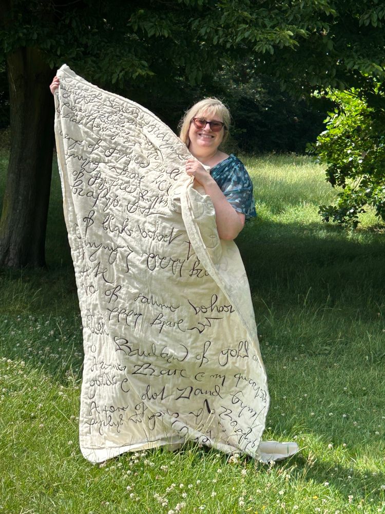 A white woman holding up a 6ft quilted piece of cream calico decorated with inked reproductions of 16th century handwriting. She is standing on grass in between trees in full leaf. 