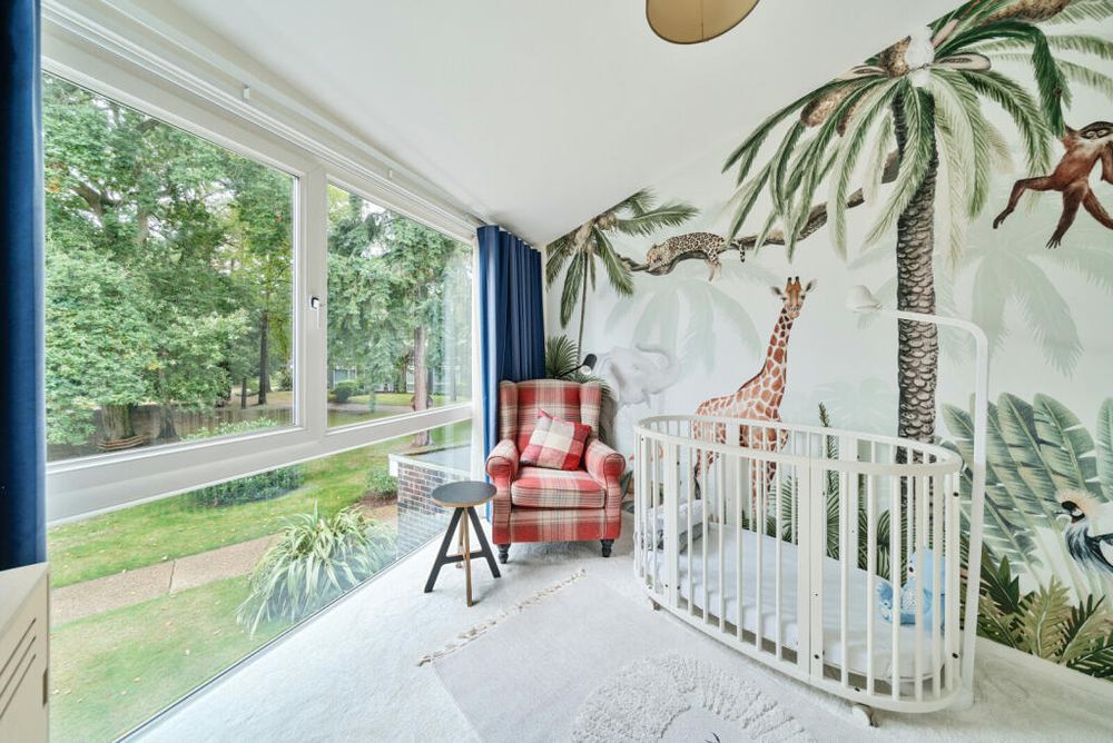One of the smaller bedrooms, still with the wall of windows. The wall next to it has a jungle mural covering it with a leopard, giraffe, monkey, and various palm tree type greenery. The furniture is a very expensive minimalist looking crib for a toddler, a wingback armchair and a stool/side table