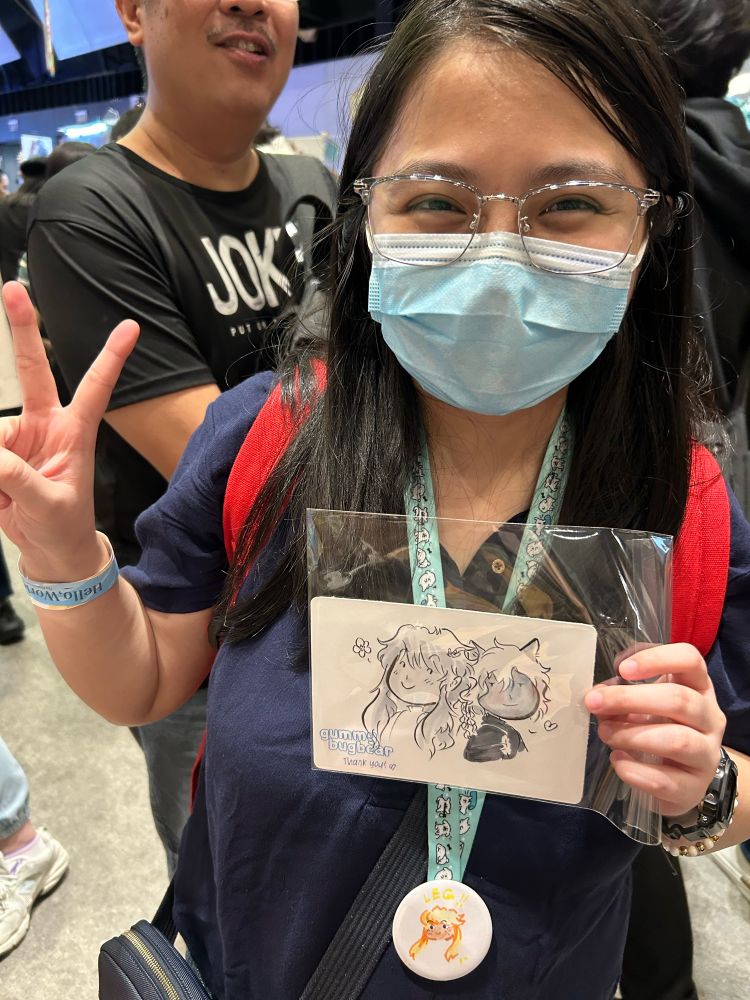 A masked congoer with long hair and glasses holding their commission and flashing a peace sign.