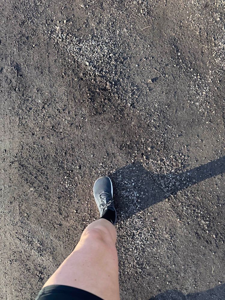 A photo of white person’s leg walking with shorts on and tennis shoes. The road is black gravel.