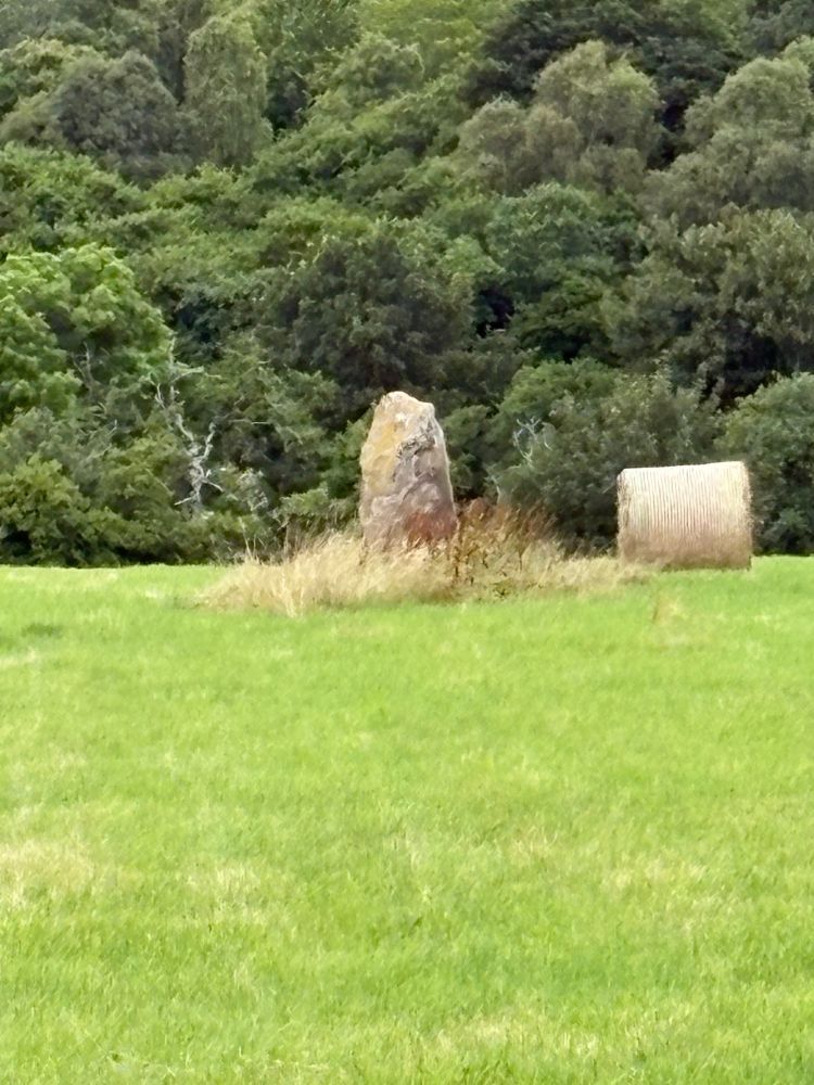 A standing stone in a hay field. There is a circular bale of hay next to it.