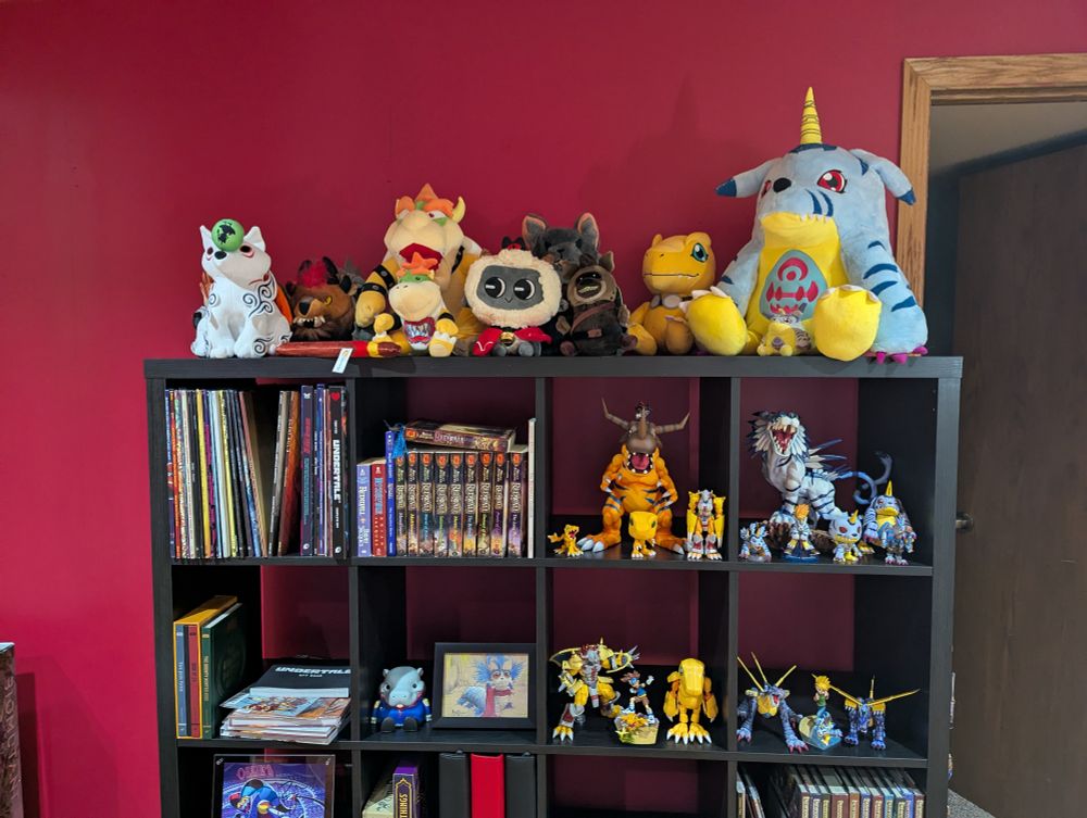 A shelf of various digimon figurines, TTRPG books, Redwall books, vinyl records, and plushes