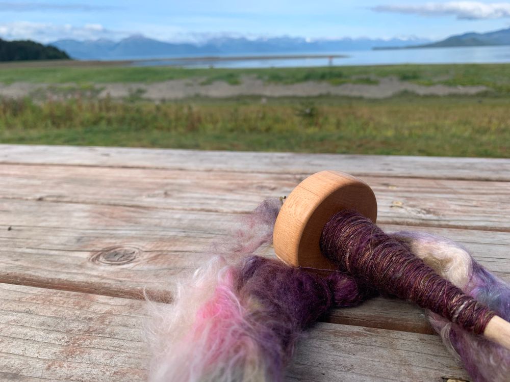 A drop spindle with a tuft of dyed wool sits on a picnic bench with a gorgeous view of the of the ocean and mountains. Alaska at its finest. 
