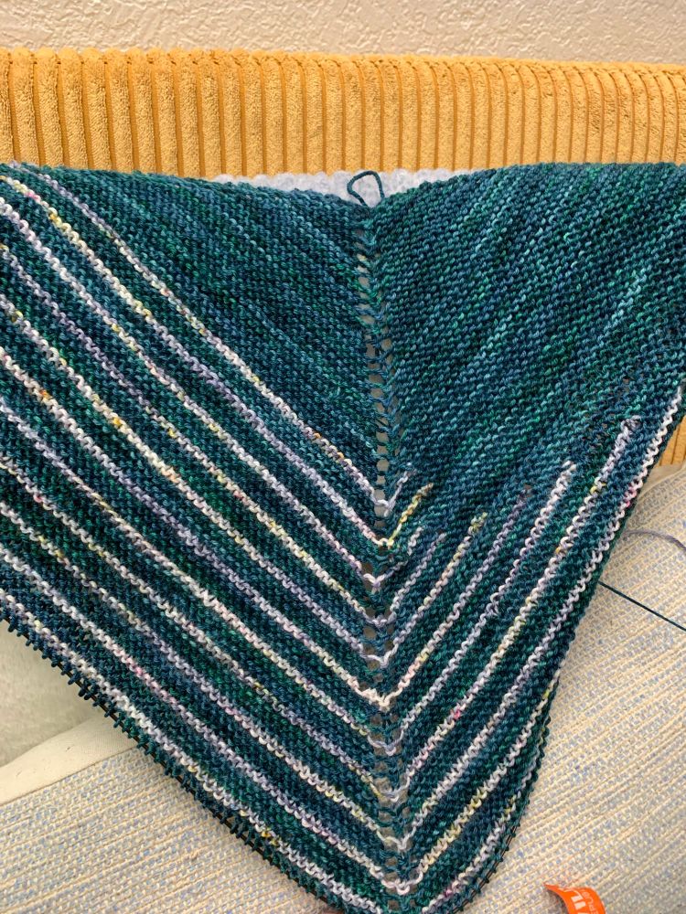 A shawl in progress, with a hand dyed teal yarn as the main color with stripes that have speckles of yellow, pink and blue.