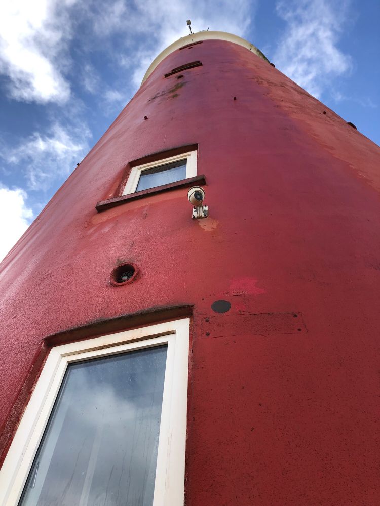 Roter Lechtturm vor blauem Himmel. Von unten und aus nächster Nähe aufgenommen, wirkt er enorm klobig.