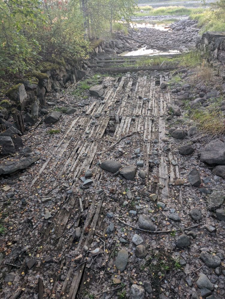 Kuva joen pohjasta kun vesi on poissa. Jokin kalanpyydyksen raunio, pitkiä puisia rakenteita.