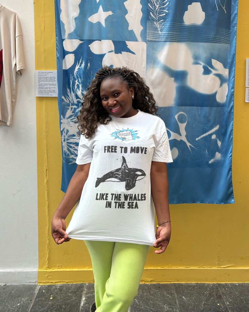 A young black woman smiling and holding the end of her t-shirt downwards. The white t-shirt reads "Free to move like whales in the sea" in black text and with a drawing of a whale in the middle.