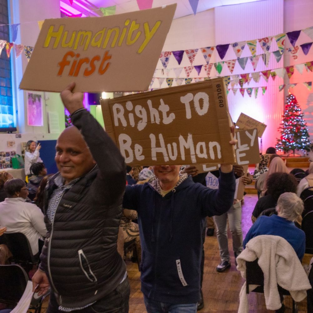 
The image depicts a vibrant community gathering inside a decorated hall. Two individuals in the foreground hold up hand-made cardboard signs. The sign on the left reads "Humanity first," while the sign on the right says "Right to be Human." The atmosphere is warm and festive, with colorful bunting hanging across the ceiling and a lit Christmas tree visible in the background. People are seated in chairs, engaged and facing the front of the hall, where the event appears to be taking place.
