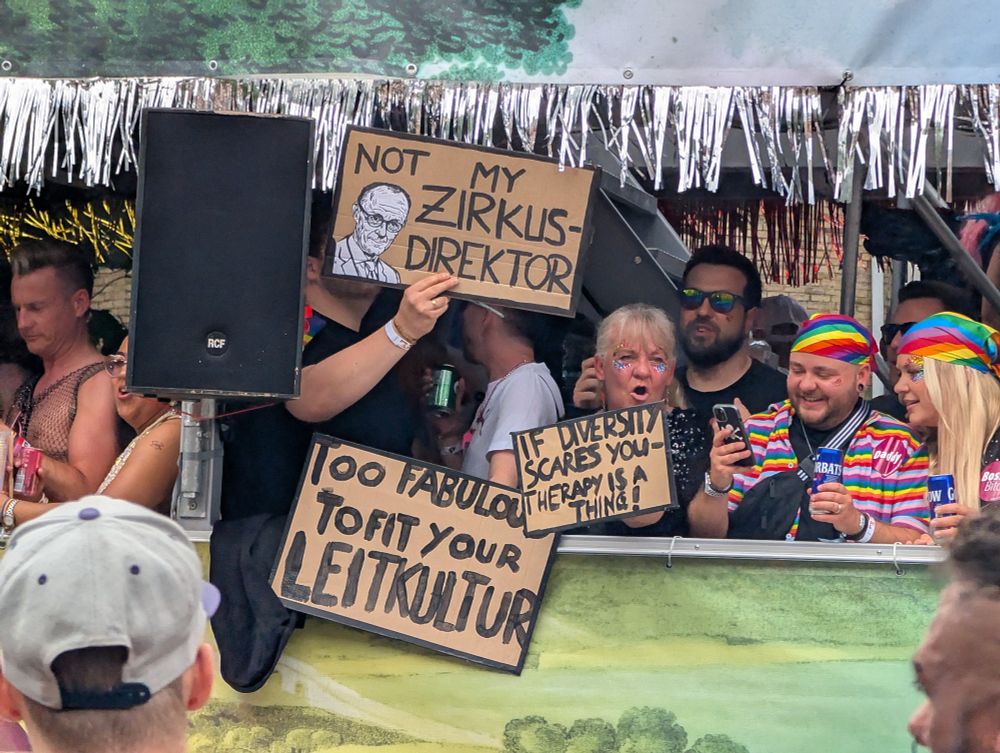 Das Bild zeigt eine fröhliche und bunte Szene auf einem Umzugswagen bei einer Pride-Parade. Der Wagen ist geschmückt mit silbernen Lametta-Girlanden und einem großflächigen Plakat mit Waldmotiv am unteren Rand. Viele Menschen stehen dicht an dicht auf dem Wagen, sie lachen, trinken und feiern. Die Stimmung wirkt ausgelassen und fröhlich.

Mehrere Personen halten selbstgemachte Schilder hoch:

Ein Schild zeigt eine gezeichnete ältere männliche Person (möglicherweise ein Politiker) und den Text: „NOT MY ZIRKUSDIREKTOR“ (eine Abwandlung des Ausdrucks „Nicht mein Zirkusdirektor“, was sinngemäß bedeutet: „Der gehört nicht zu uns“).

Ein weiteres Schild sagt: „100 FABULOUS TO FIT YOUR LEITKULTUR“ (frei übersetzt: „Zu fabelhaft, um in eure Leitkultur zu passen“).

Ein drittes Schild trägt die Botschaft: „IF DIVERSITY SCARES YOU – THERAPY IS A THING!“ (zu Deutsch: „Wenn Vielfalt dir Angst macht – es gibt so etwas wie Therapie!“).