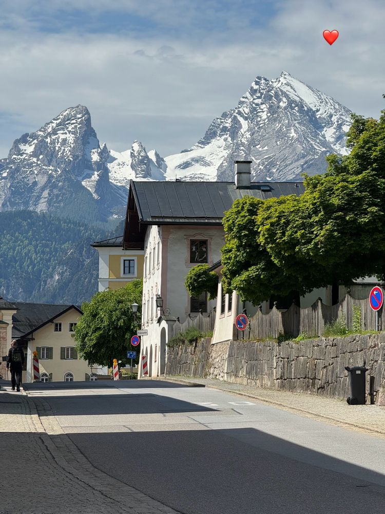 Blick auf den Watzmann
Im Vordergrund eine Straße die leicht Bergauf führt und 2 dunkle Schatten von Häusern zeigt. Rechts eine graue Steinmauer auf der ein grauer Holzzaun steht der wellenartig geformt ist und aus dem kleine, grüne Baumkronen herausragen. Danach kommt ein weises Haus mit gelb eingefassten Fenstern und dunklen Dach. Angrenzend sieht man noch einen Teil von einer gelben Hausfassade mit weißen Absetzungen. Zwischen diesen Häusern steht vorne ein grüner Baum.
Geradeaus befindet sich wieder ein gelbes Haus mit grünen Fensterläden.
Über dem Haus ist ein großes Waldstück. Jetzt erstreckt sich
Majestätisch der Berg Watzmann über die ganze Breite des Bildes  der teilweise mit Schnee bedeckt ist. Der linke Teil des Berges wird die 
Mutter genannt in der Mitte befinden sich die Kinder (2kleine Bergspitzen) und rechts der dominante Vater die größte Bergspitze.
Der Himmel etwas nebelig wird nach oben hin blau.