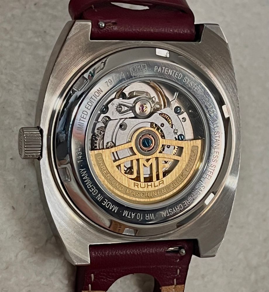 Das Bild zeigt die Rückseite einer Uhr mit einer transparenten Abdeckung, die die mechanische Uhrwerkstruktur sichtbar macht. Die Uhrwerkkomponenten sind silberfarben, der Automatikrotor ist in Gold gearbeitet. Die Komponenten sind sorgfältig gearbeitet und zeigen die Präzision und Komplexität mechanischer Uhren. 