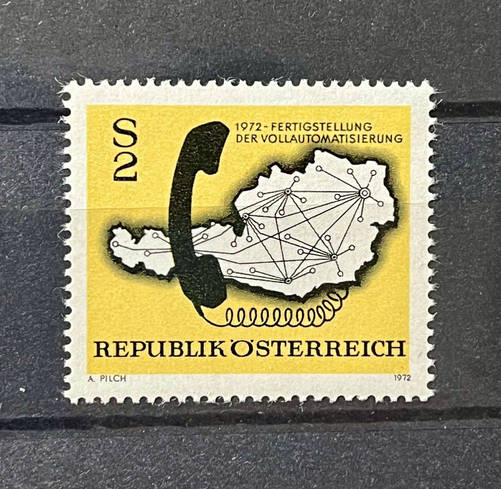 Das Bild zeigt eine österreichische Briefmarke aus dem Jahr 1972. Die Marke hat einen gelben Hintergrund und zeigt die Silhouette Österreichs in Grau. Auf der Marke ist ein schwarzes Telefonhörer abgebildet, das mit einem Netzwerk von Linien und Punkten verbunden ist, das die gesamte Landesgrenze bedeckt. Diese Darstellung symbolisiert die vollständige Automatisierung der Telekommunikation in Österreich im Jahr 1972. Oben auf der Marke steht in schwarzer Schrift 
