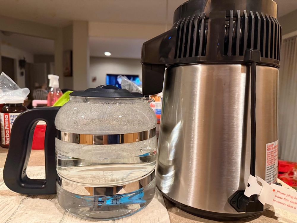 The photo shows a stainless steel water distiller with a black top next to a glass carafe filled with water. The carafe has a black handle and a black lid. The distiller is plugged in and sits on a kitchen countertop.