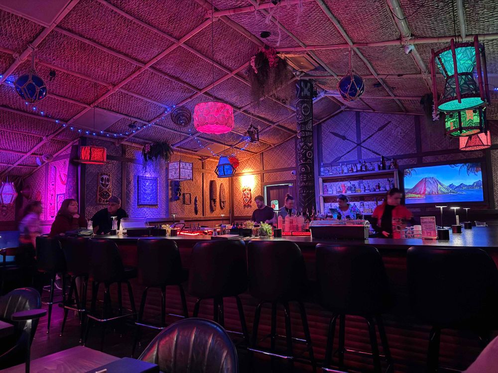 This photo depicts the interior of a tiki-themed bar or lounge. The setting features vibrant, colorful lighting with hues of pink, purple, and blue, creating a lively and tropical atmosphere. The ceiling is adorned with woven bamboo or straw-like materials, and various decorative hanging lanterns in different shapes and colors add to the exotic ambiance. Behind the bar, bottles of liquor are neatly arranged on shelves, and a large, vivid screen of a tropical island scene with mountains and ocean is prominently displayed. Patrons are seated at the bar and tables, while bartenders are busy preparing drinks. The overall vibe suggests a fun, relaxed environment with a tropical, island-inspired decor.