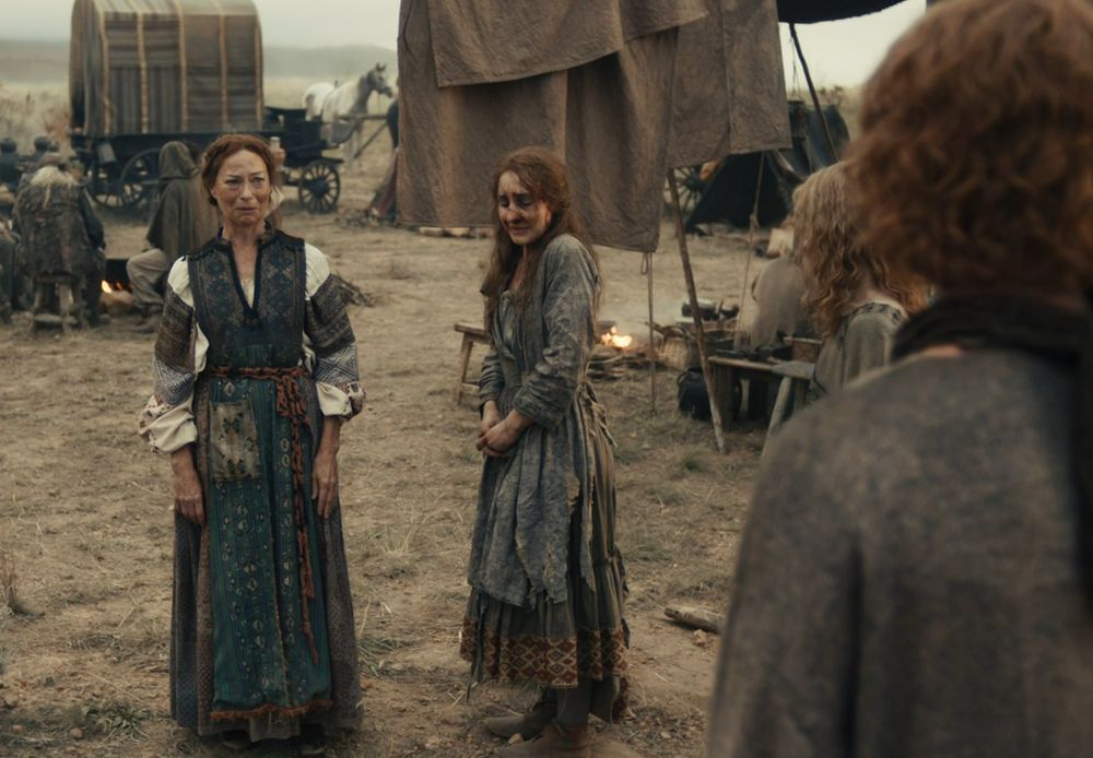 An upset white woman and her daughter stand amidst a wagon camp. they're wearing travel-worn dresses, and the daughter's face is heavily bruised.  We see the back of Lewin, a young man with red wavy hair.