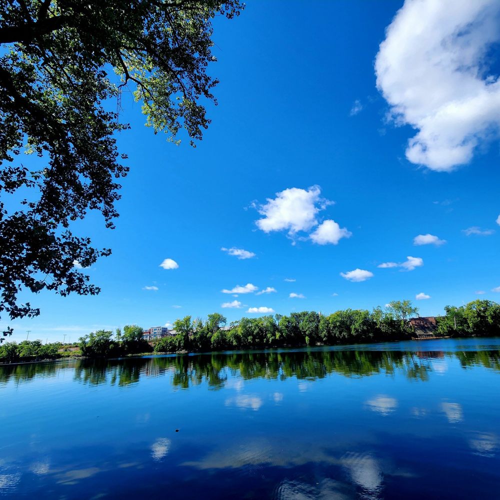 Blue sky and little white clouds reflected in a blue river surrounded by trees.