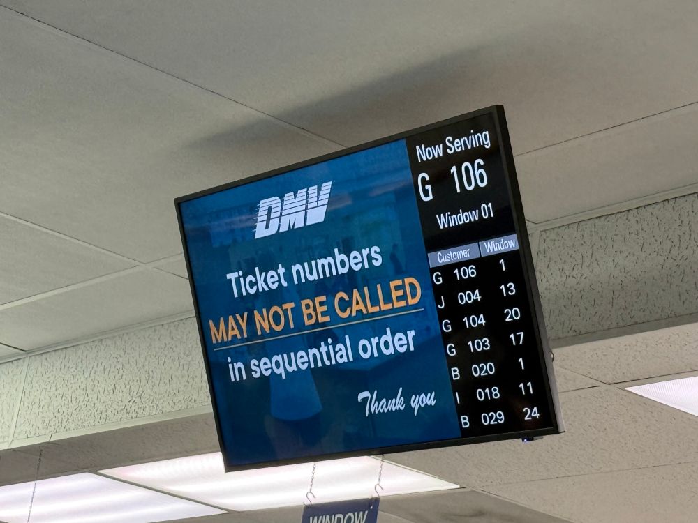 an elevated ceiling-mounted ticket number screen at the DMV saying ticket numbers may not be called in sequential order