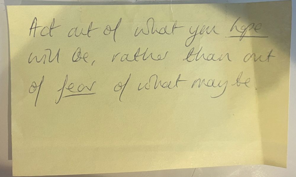 Pale yellow post-it note stuck to a wall that reads in joined-up writing “act out of what you hope will be, rather than out of fear of what may be”.
