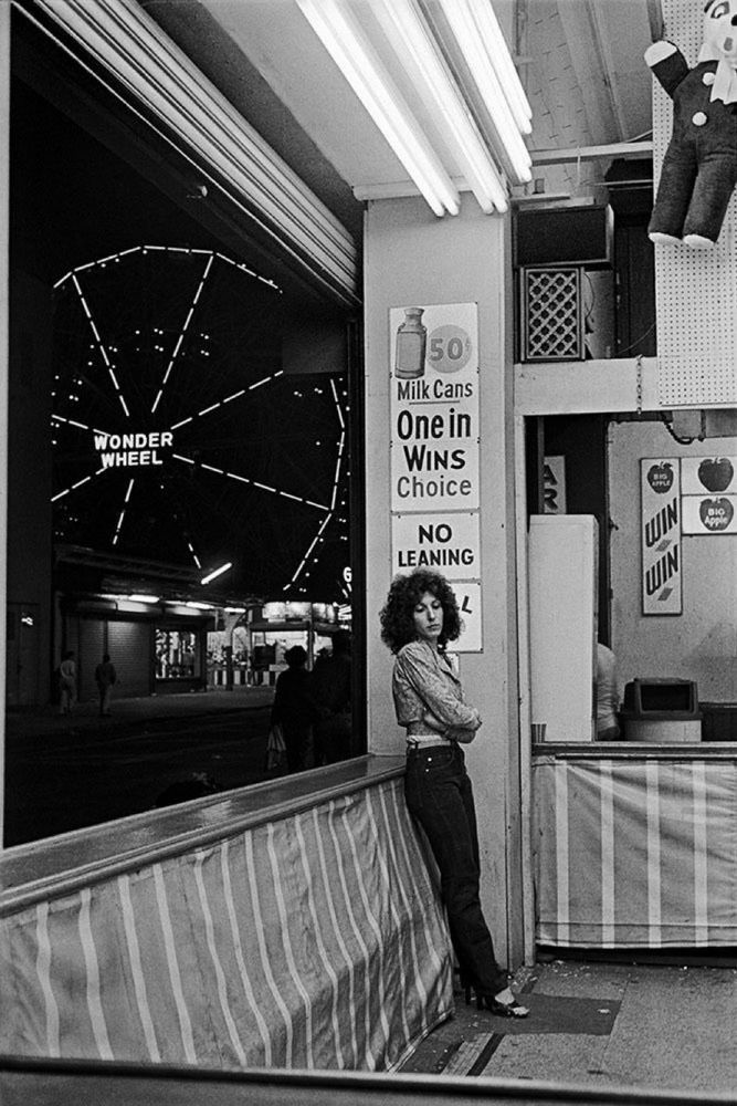 A black and white photo of a woman at a fairground stand in Coney Island in the 1970s, who has been captured by photographer Hazel Hankin leaning against a counter above which is a sign that says “no leaning. A ferris wheel is lit up in the background. The image was shared on BlueSky by @desdelboy.bsky.social