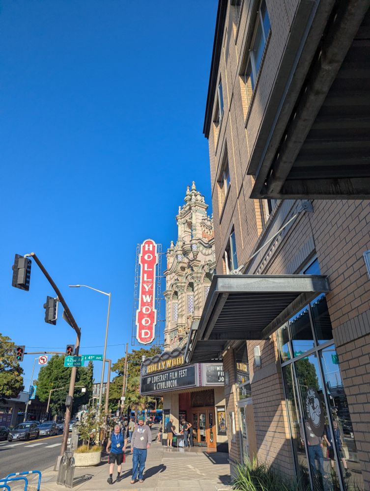 A view of Portland, Oregon's Hollywood Theater peeking out from behind a beige-colored contemporary building.  It is a gorgeous 1926 movie house with an ornate baroque facade and a large red vertical sign with "Hollywood" in illuminated white letters.  The marquee says "HP Lovecraft Film Fest & Cthulhucon."  Several people with film festival lanyards mill about.