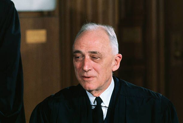 A photograph of justice Harlan.  He is an old white man with thinning silver hair, a thin face, and a black tie on under his judicial robe.  He is squinting a bit and his mouth is slightly open, like he is in the middle of saying "oh my."