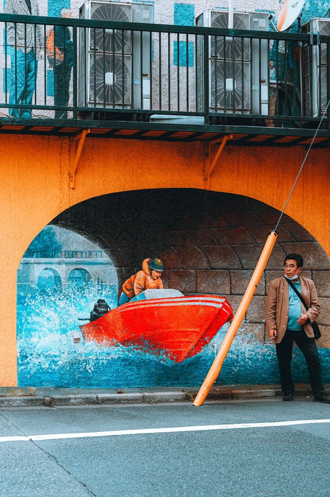 Photo d’un home fumant une cigarette devant une fresque street art, représentant un bateau à moteur passant sous un pont