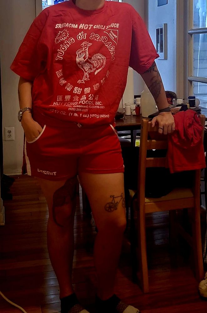 Riley in red siracha shirt and red 70s style work out shorts, leaning on a chair
