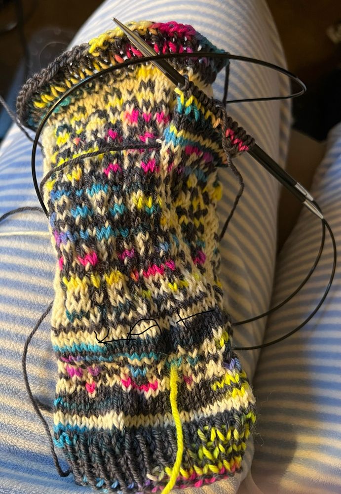 Knitting fingerless mittens with circular knitting needles finishing up the thump. Multicolored stitches of pink, blue, black, yellow and white. 