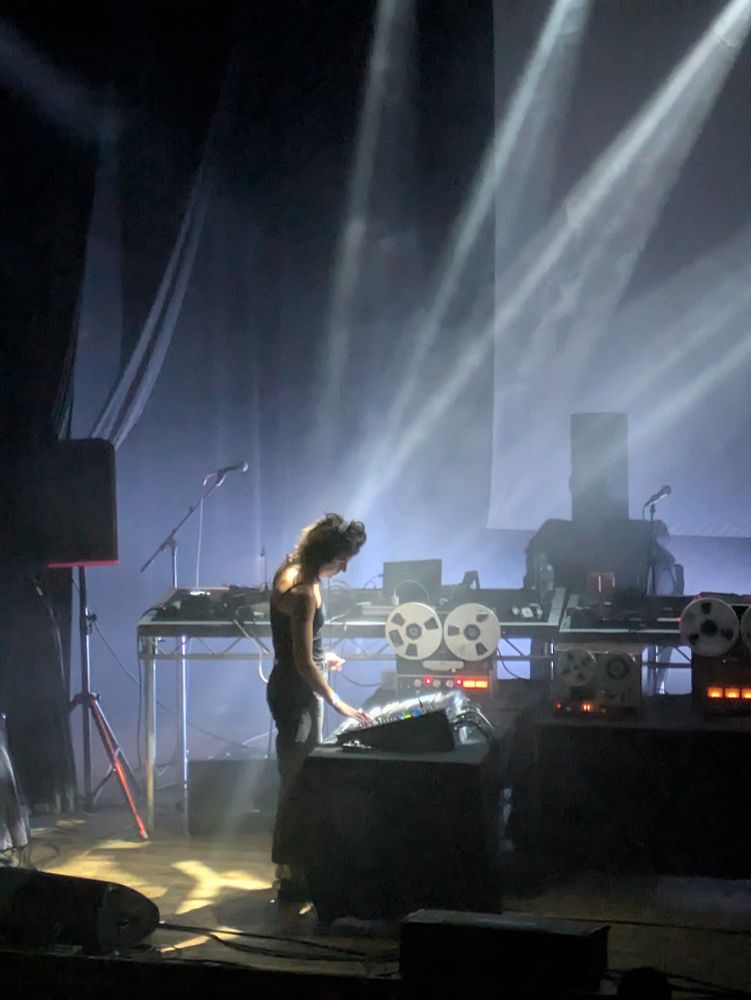 A woman stands on stage lit by spotlights. Old fashioned tape machines surround her.
