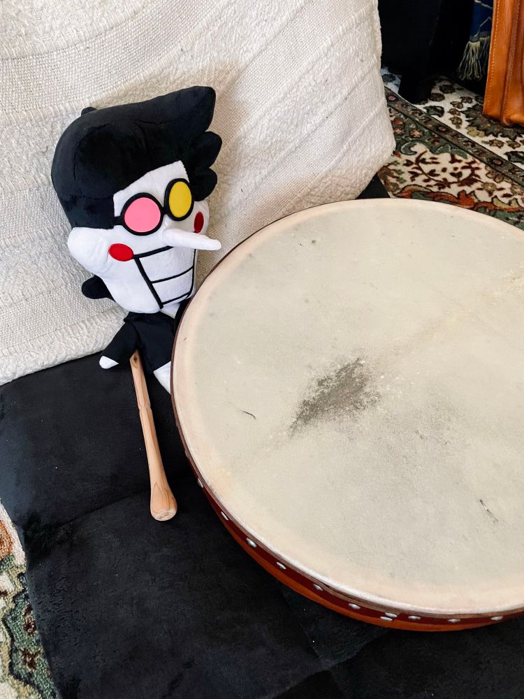A plush doll of Spamton placed on a floor chair. A bodhrán is resting over top him and a beater happens to be tucked under his tiny little arm. It is all much too big for him to play properly.