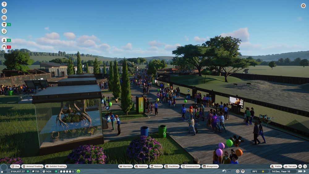 Captura de pantalla de un juego en que se ve un zoo con varios recintos, mucha gente (varies de elles con globos) y vegetación muy variada. En la parte de abajo y en la superior izquierda se ve una UI de juego de gestión.